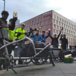 Vor dem Dortmunder U stehen sieben Personen in Fahrradkleidung mit Lastenfahrrädern und winken freundlich in die Kamera